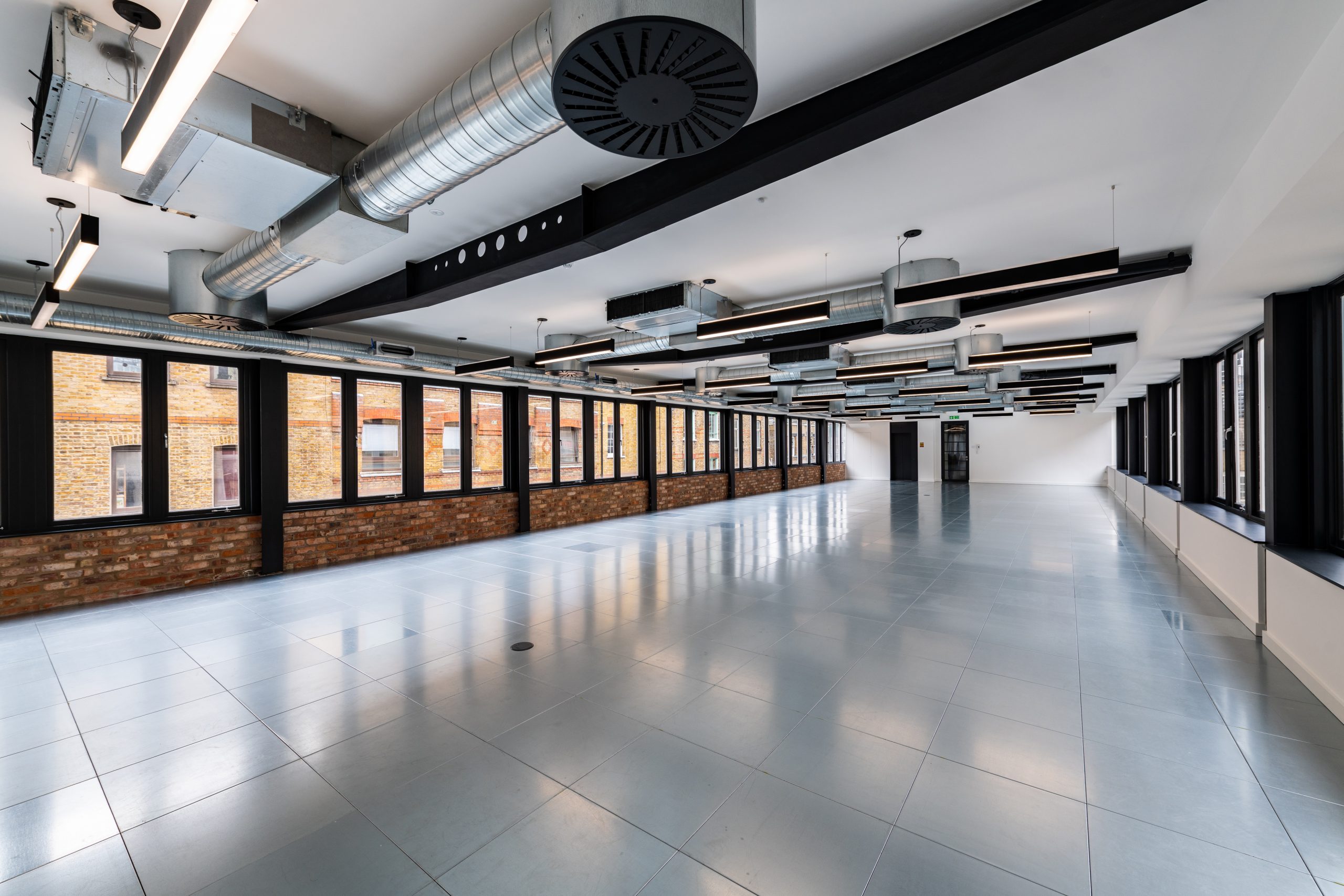 grey tiled floor, low white ceilings, bright lighting ,industrial ducting, wall of windows with black frame and the lower quarter is a brick wall